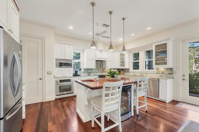 kitchen featuring decorative light fixtures, white cabinets, beverage cooler, stainless steel appliances, and a center island with sink