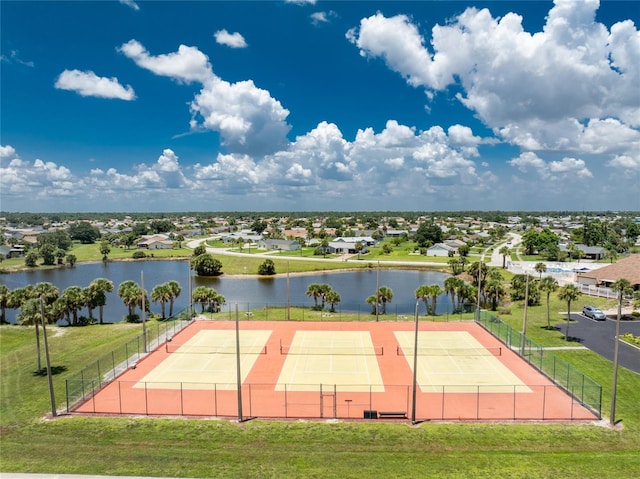 view of tennis court with a water view, a residential view, fence, and a lawn