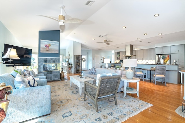 living room with ceiling fan, a tiled fireplace, high vaulted ceiling, and light wood-type flooring