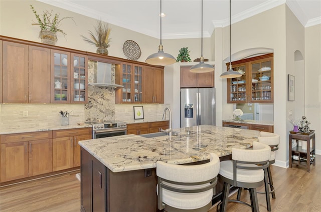 kitchen with sink, a center island with sink, hanging light fixtures, stainless steel appliances, and wall chimney range hood