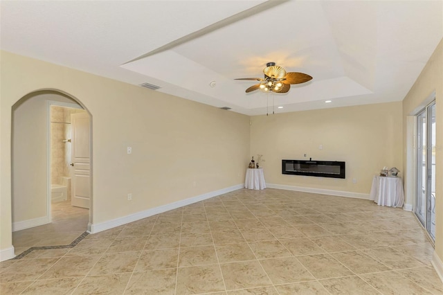 unfurnished living room with baseboards, arched walkways, a ceiling fan, a glass covered fireplace, and a tray ceiling