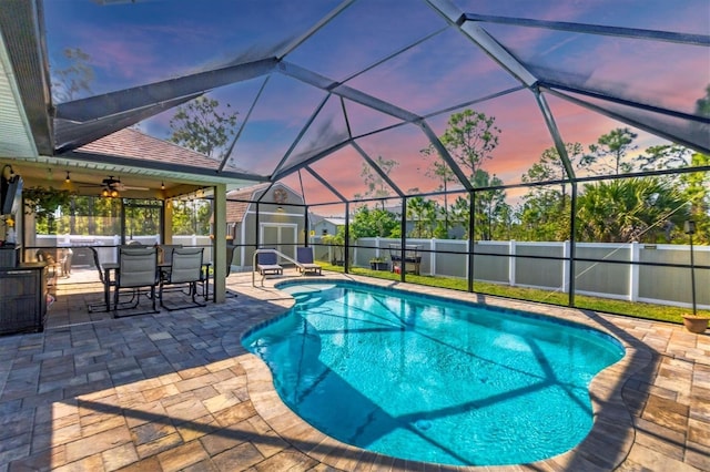 pool at dusk with a patio, a lanai, ceiling fan, and a shed