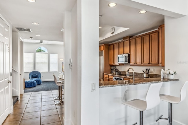 kitchen with light tile patterned floors, a breakfast bar area, appliances with stainless steel finishes, dark stone countertops, and kitchen peninsula