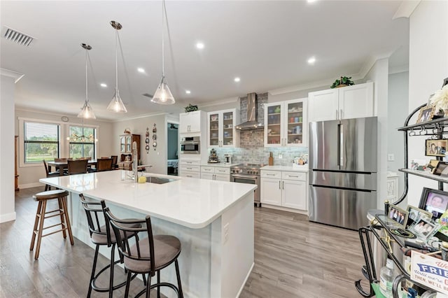 kitchen featuring a kitchen island with sink, stainless steel appliances, white cabinets, wall chimney range hood, and pendant lighting