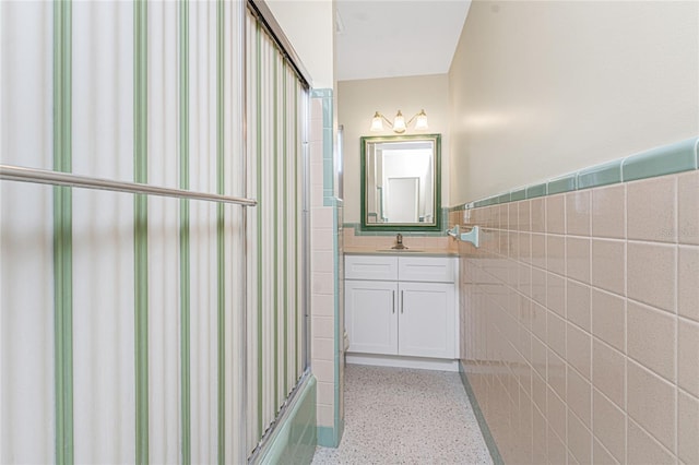 bathroom featuring a wainscoted wall, speckled floor, shower / bath combination with glass door, vanity, and tile walls