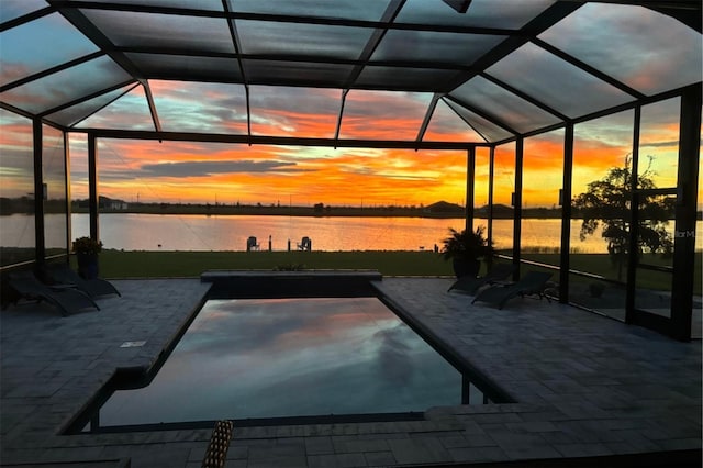 pool at dusk with glass enclosure, a patio, and a water view