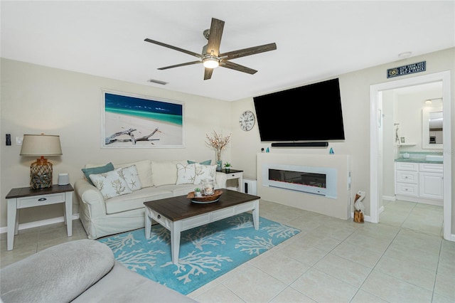 living room featuring light tile patterned floors, ceiling fan, visible vents, baseboards, and a glass covered fireplace