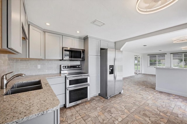 kitchen with a sink, light stone countertops, gray cabinets, stainless steel appliances, and backsplash