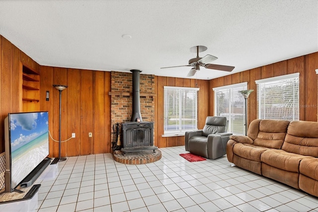 living room with light tile patterned floors, wooden walls, a ceiling fan, a wood stove, and a textured ceiling