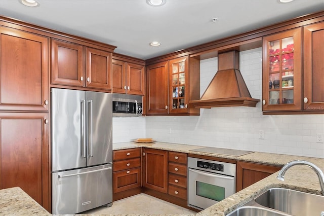 kitchen with stainless steel appliances, premium range hood, a sink, decorative backsplash, and glass insert cabinets