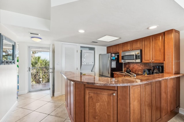 kitchen featuring stone countertops, stacked washer and dryer, appliances with stainless steel finishes, brown cabinets, and decorative backsplash