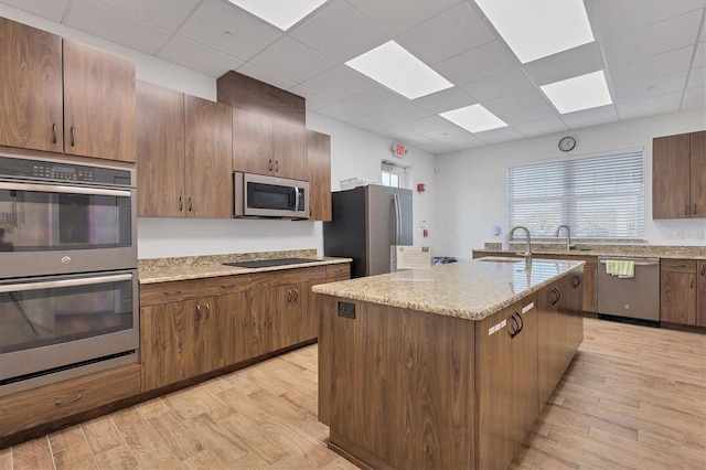 kitchen featuring stainless steel appliances, a center island with sink, light wood-style flooring, and light stone countertops