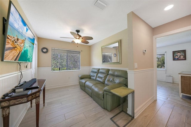 living area featuring light wood-style flooring, visible vents, a wainscoted wall, and ceiling fan