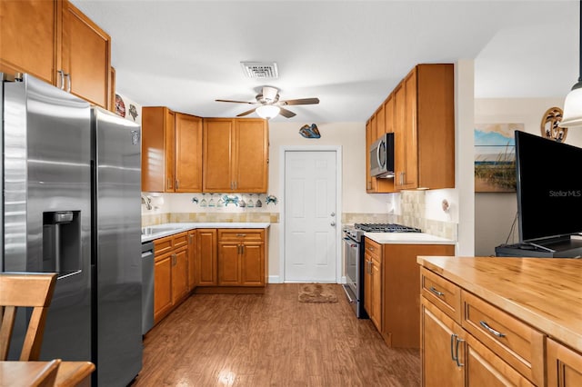 kitchen with a sink, brown cabinetry, stainless steel appliances, a ceiling fan, and dark wood-style flooring