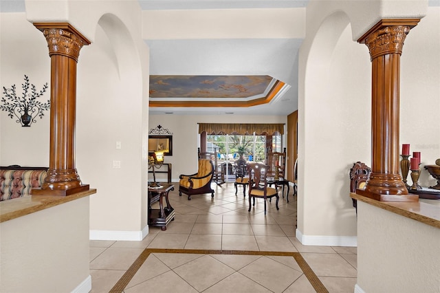 hall with a raised ceiling, light tile patterned floors, baseboards, and ornate columns