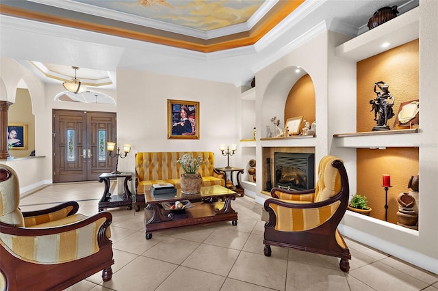 tiled living room featuring a tray ceiling, decorative columns, arched walkways, ornamental molding, and a glass covered fireplace