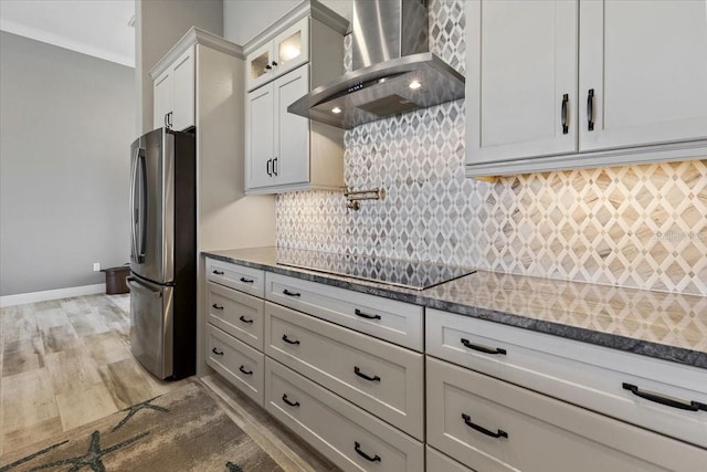 kitchen featuring black electric cooktop, light wood-style flooring, wall chimney exhaust hood, freestanding refrigerator, and backsplash