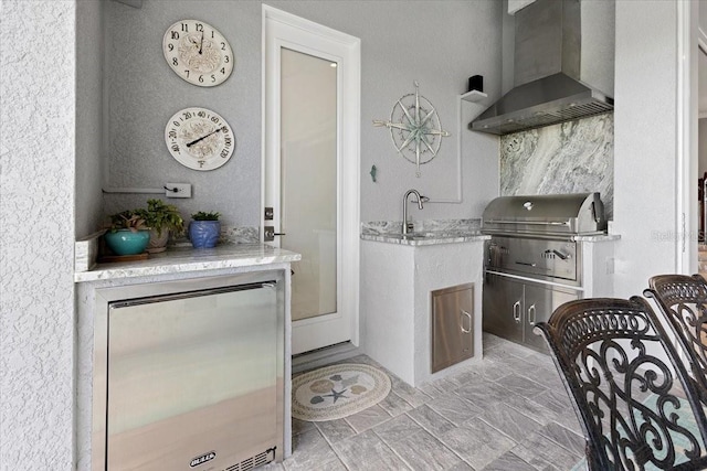 kitchen featuring wall chimney range hood, fridge, a textured wall, and light stone counters