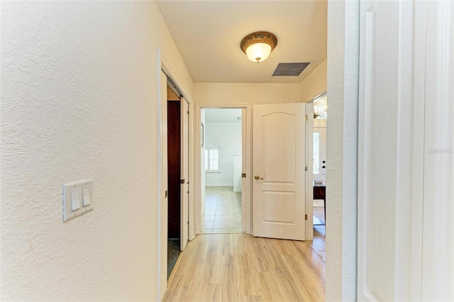 hallway featuring a textured wall, light wood-type flooring, and visible vents