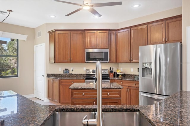 kitchen featuring appliances with stainless steel finishes, a ceiling fan, brown cabinetry, dark stone countertops, and recessed lighting