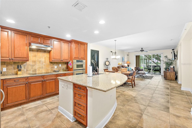 kitchen with decorative light fixtures, backsplash, a kitchen island with sink, black electric stovetop, and light stone countertops