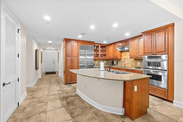 kitchen featuring appliances with stainless steel finishes, tasteful backsplash, sink, light stone counters, and a center island with sink