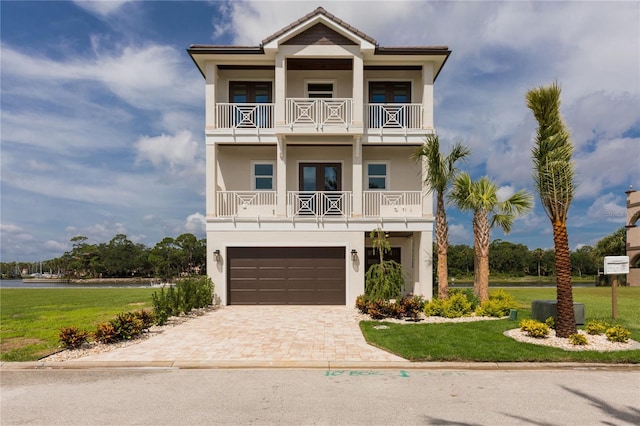 coastal inspired home with a balcony, a front lawn, decorative driveway, and stucco siding