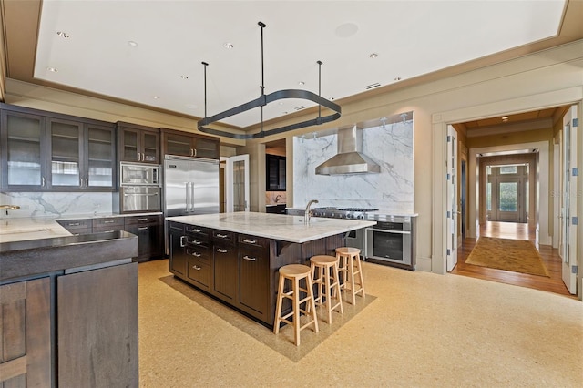 kitchen with wall chimney exhaust hood, tasteful backsplash, a center island with sink, and dark brown cabinetry