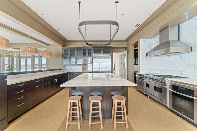 kitchen with pendant lighting, a kitchen island with sink, backsplash, wall chimney range hood, and built in appliances