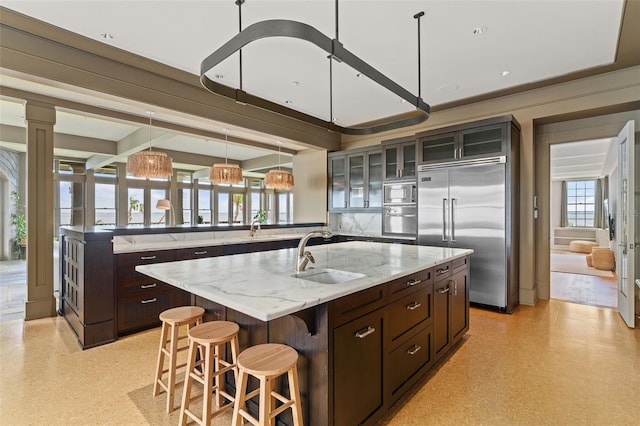 kitchen with an island with sink, light stone countertops, sink, pendant lighting, and built in appliances