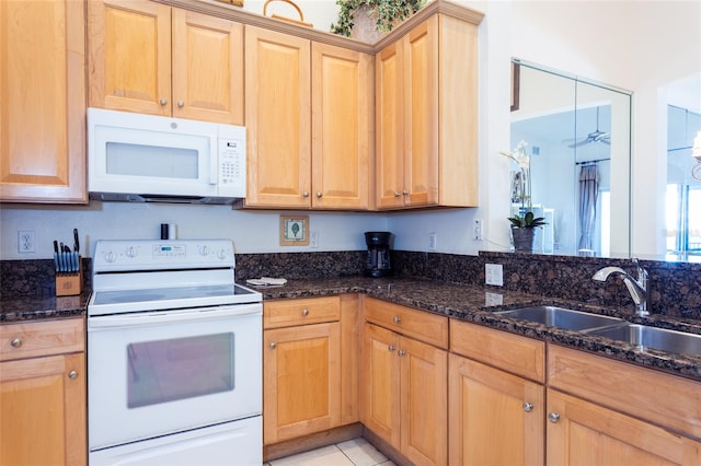 kitchen featuring white appliances, light tile patterned floors, light brown cabinets, dark stone counters, and a sink