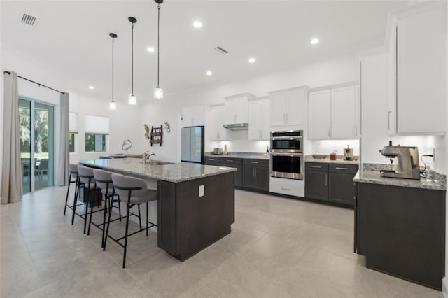 kitchen featuring a center island with sink, pendant lighting, stainless steel appliances, light stone countertops, and light tile flooring