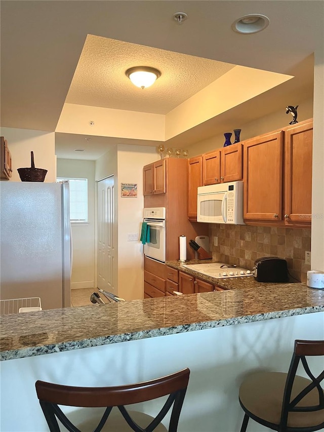 kitchen featuring a breakfast bar area, backsplash, light stone counters, kitchen peninsula, and white appliances