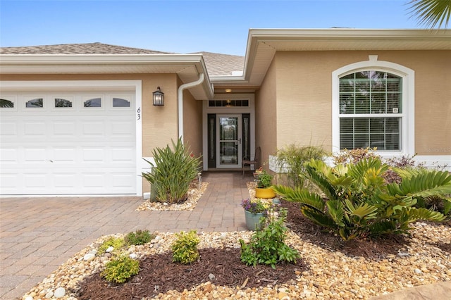 doorway to property with decorative driveway, roof with shingles, an attached garage, and stucco siding