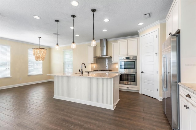 kitchen with wall chimney range hood, stainless steel appliances, a kitchen island with sink, and hanging light fixtures