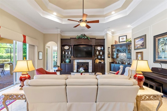 tiled living room with ornamental molding, ceiling fan, a raised ceiling, and a fireplace