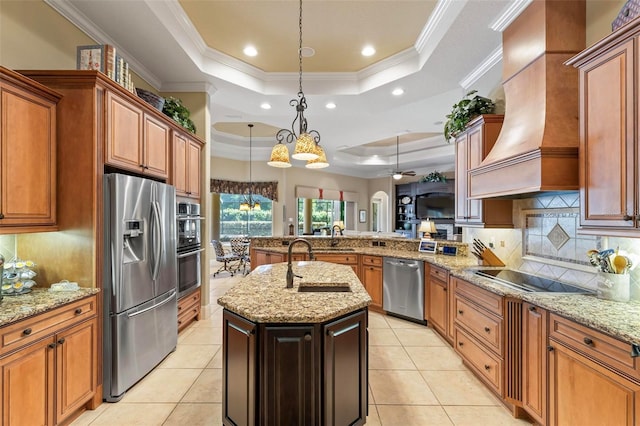 kitchen with custom exhaust hood, appliances with stainless steel finishes, a raised ceiling, pendant lighting, and a kitchen island with sink