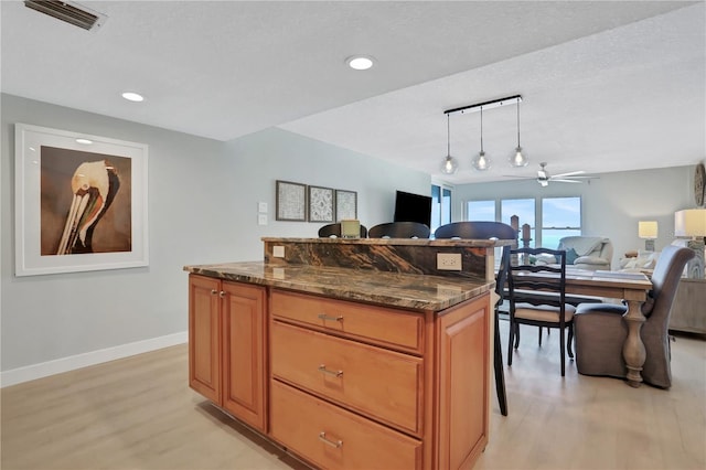 kitchen with ceiling fan, a kitchen island, light wood-type flooring, dark stone countertops, and pendant lighting