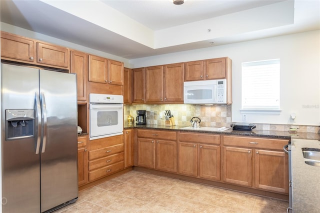 kitchen featuring white appliances, a raised ceiling, dark stone countertops, and backsplash