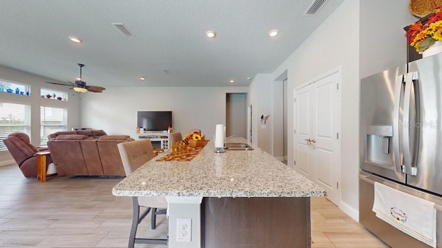 kitchen featuring ceiling fan, sink, stainless steel fridge with ice dispenser, light stone counters, and a kitchen island with sink