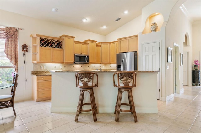 kitchen with backsplash, vaulted ceiling, appliances with stainless steel finishes, light brown cabinetry, and dark stone counters