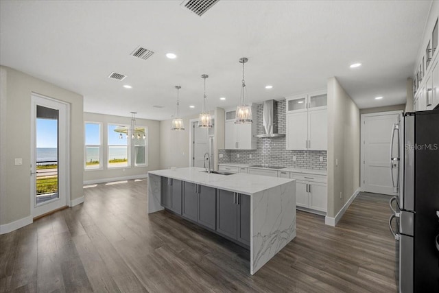 kitchen with wall chimney range hood, stainless steel fridge, white cabinets, and a center island with sink