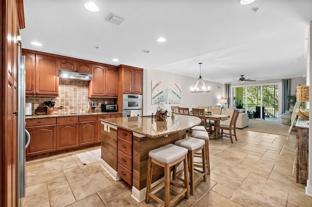 kitchen with decorative light fixtures, backsplash, a kitchen island, double oven, and light stone counters