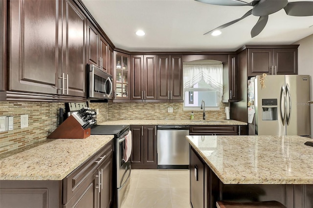 kitchen with light stone counters, stainless steel appliances, dark brown cabinets, ceiling fan, and sink