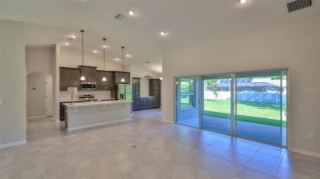 kitchen featuring appliances with stainless steel finishes, high vaulted ceiling, tasteful backsplash, an island with sink, and decorative light fixtures