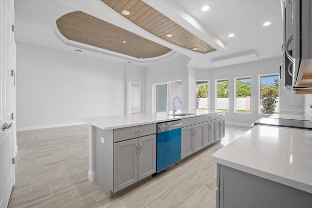 kitchen with a center island with sink, gray cabinets, sink, stainless steel dishwasher, and a raised ceiling