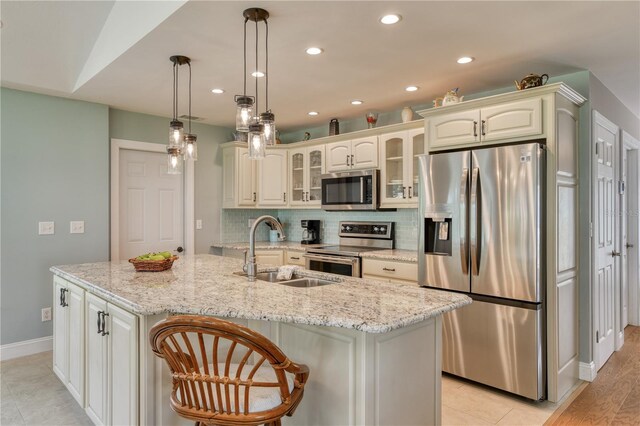 kitchen with a center island with sink, a sink, backsplash, appliances with stainless steel finishes, and glass insert cabinets