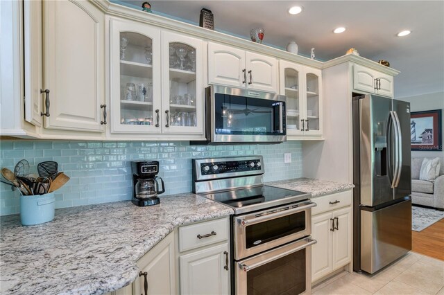 kitchen with backsplash, stainless steel appliances, light tile patterned floors, glass insert cabinets, and light stone countertops