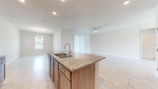 kitchen with sink, ceiling fan with notable chandelier, pendant lighting, light stone counters, and a kitchen island with sink