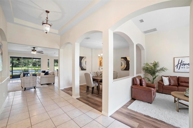 living room with a raised ceiling, light tile patterned floors, and ceiling fan with notable chandelier